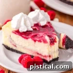 slice of white chocolate raspberry cheesecake on a round white plate topped with whipped cream. three raspberries are sitting next to the cheesecake on the plate.