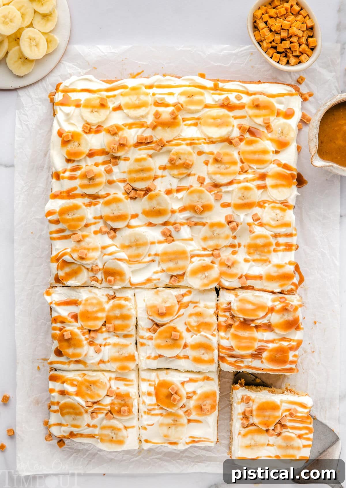 Dreamy Banoffee Poke Cake 3 A top-down view of a whole Banoffee Poke Cake with six slices already cut, resting on white parchment paper. A bowl of sliced bananas and toffee bits sits beside the cake.