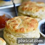 A plate of flaky, golden brown homemade biscuits, some brushed with melted butter, with a jar of jelly in the background.