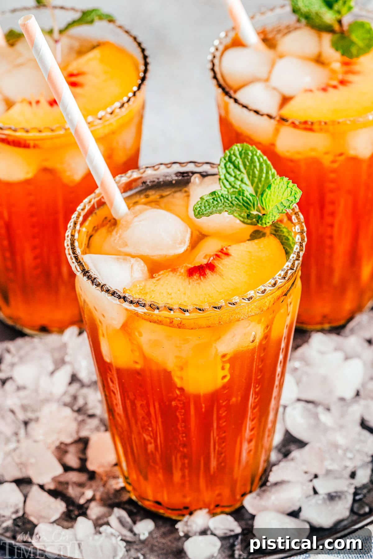 Sun-Kissed Peach Sip 2 Three tall glasses filled with homemade peach iced tea, garnished with fresh peach slices and a sprig of mint. The glasses are arranged on a tray covered with ice.