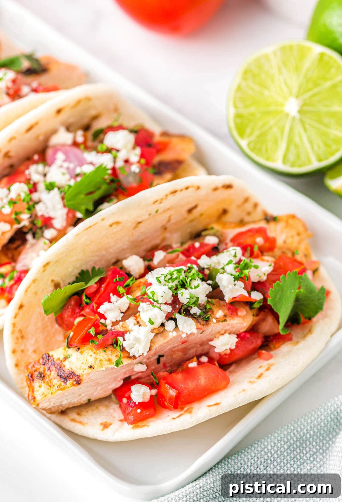 three chicken tacos on a white rectangular plate topped with pico de gallo, cilantro and queso fresco. A half a lime can be seen in the background.