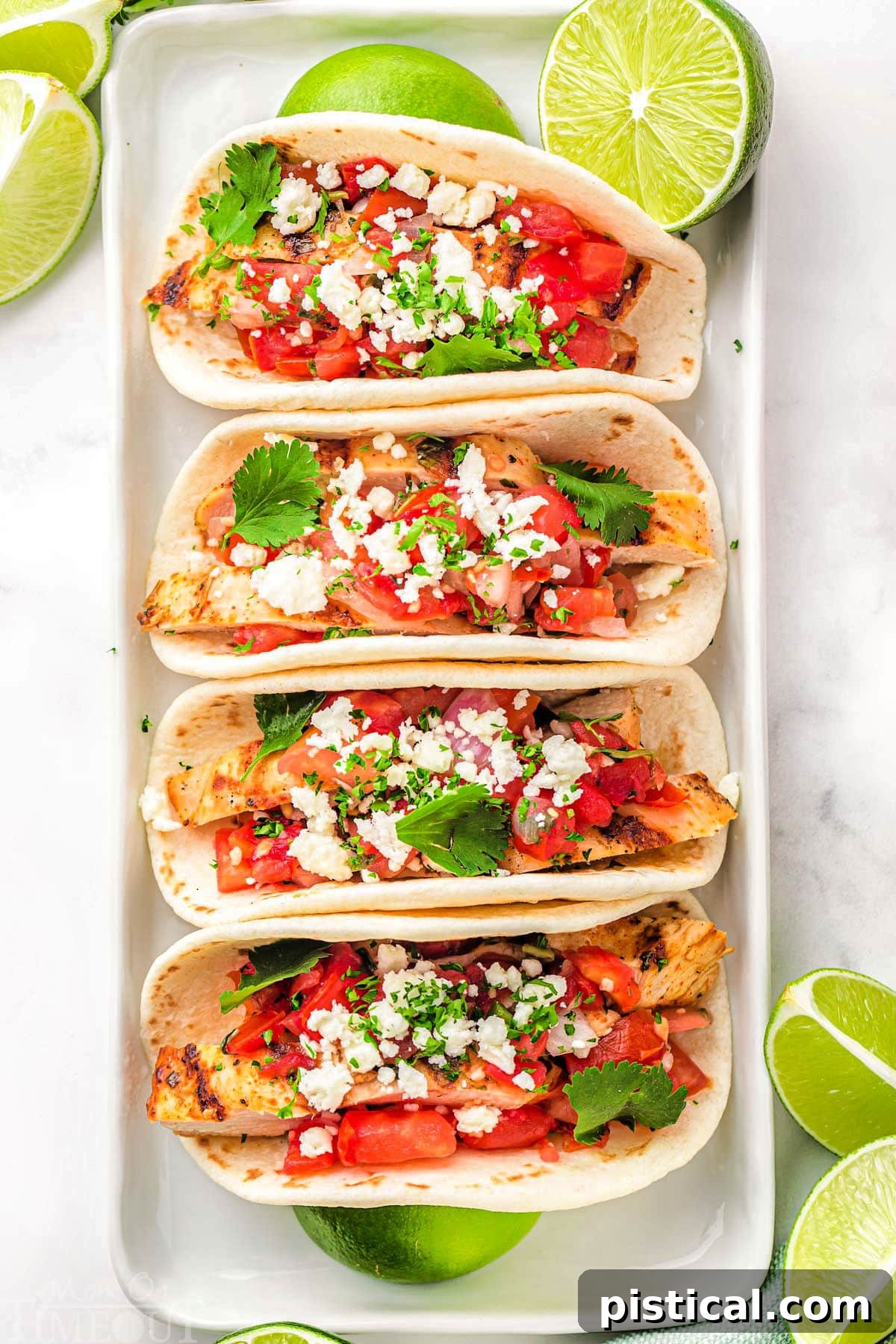 top down look at four grilled chicken tacos on a white plate topped with pico de gallo, cilantro and queso fresco. Lime wedges are scattered around the tacos.