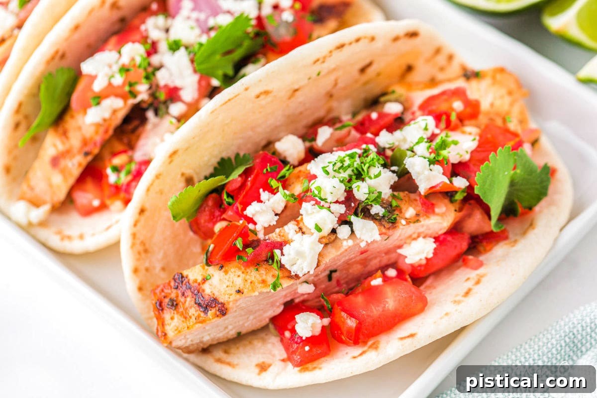 three chicken tacos on a white rectangular plate topped with pico de gallo, cilantro and queso fresco. A half a lime can be seen in the background.