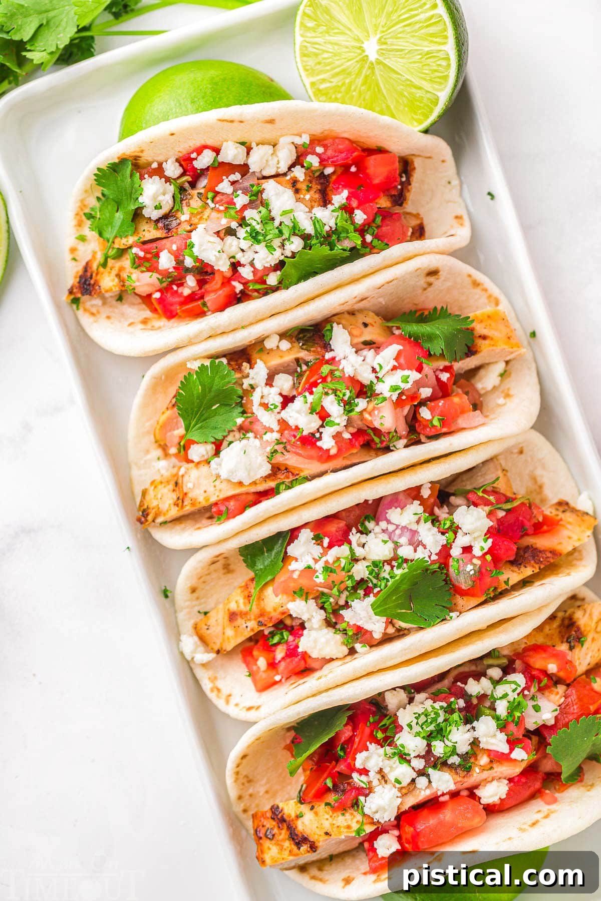 top down look at four grilled chicken tacos on a white plate topped with pico de gallo, cilantro and queso fresco. Lime wedges are scattered around the tacos.