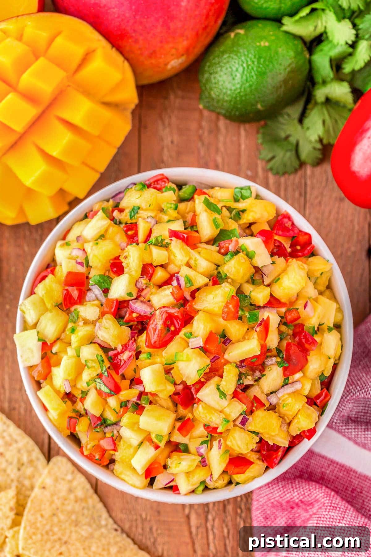top down look at pineapple mango salsa in a white bowl surround by chips, mango, limes and cilantro. 