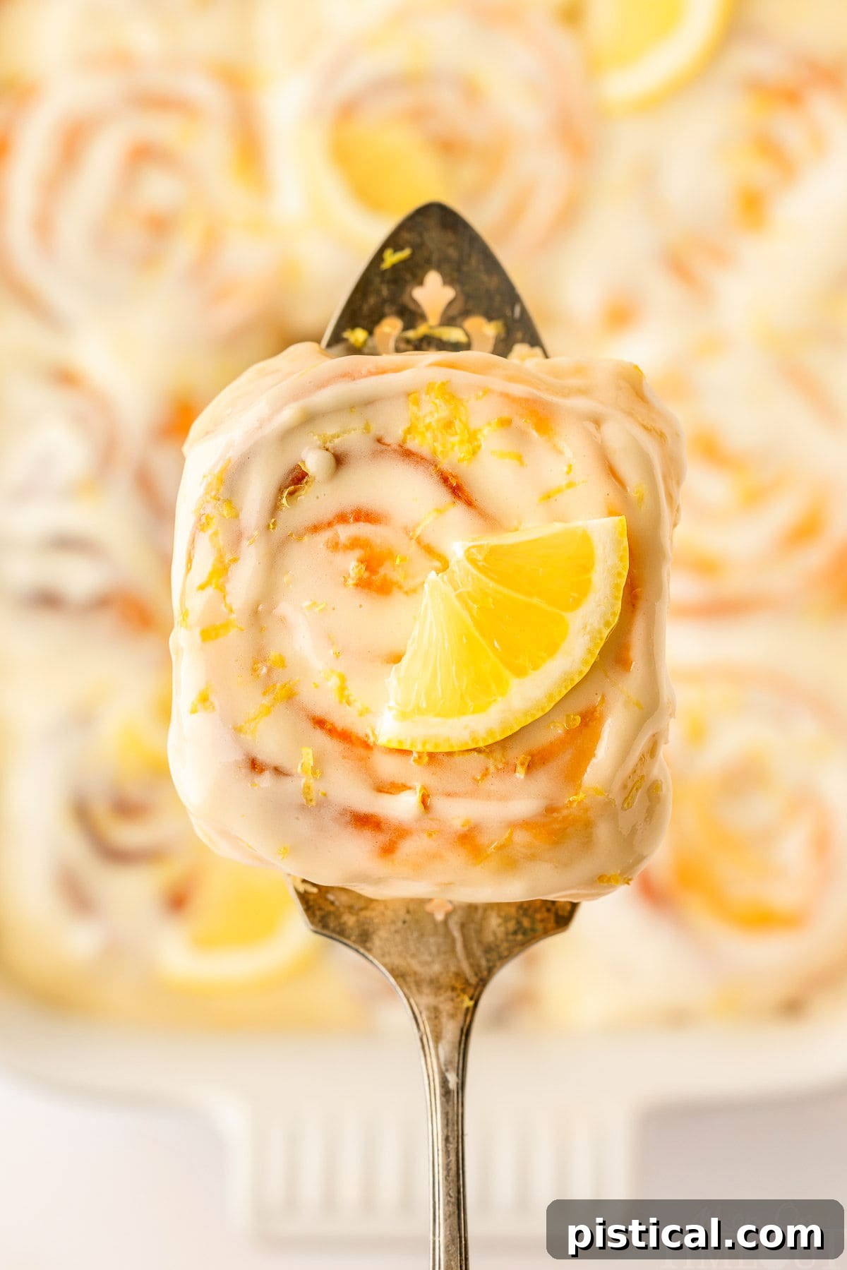 Zesty Lemon Swirls 9 A top-down view of a single lemon roll held on a spatula, just lifted from the pan of other glazed rolls.