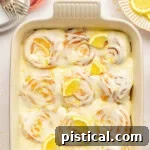 Zesty Lemon Swirls 10 An overhead view of a pan of lemon sweet rolls topped with a lemon cream cheese glaze, lemon slices and lemon zest. Baking dish is white.