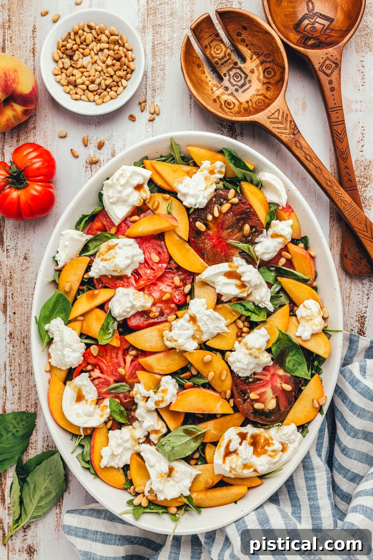 A beautifully arranged fresh Peach Burrata Salad featuring juicy peach slices, creamy burrata cheese, and vibrant heirloom tomatoes, all lightly dressed with balsamic vinaigrette and garnished with toasted pine nuts, served on a white oval platter.