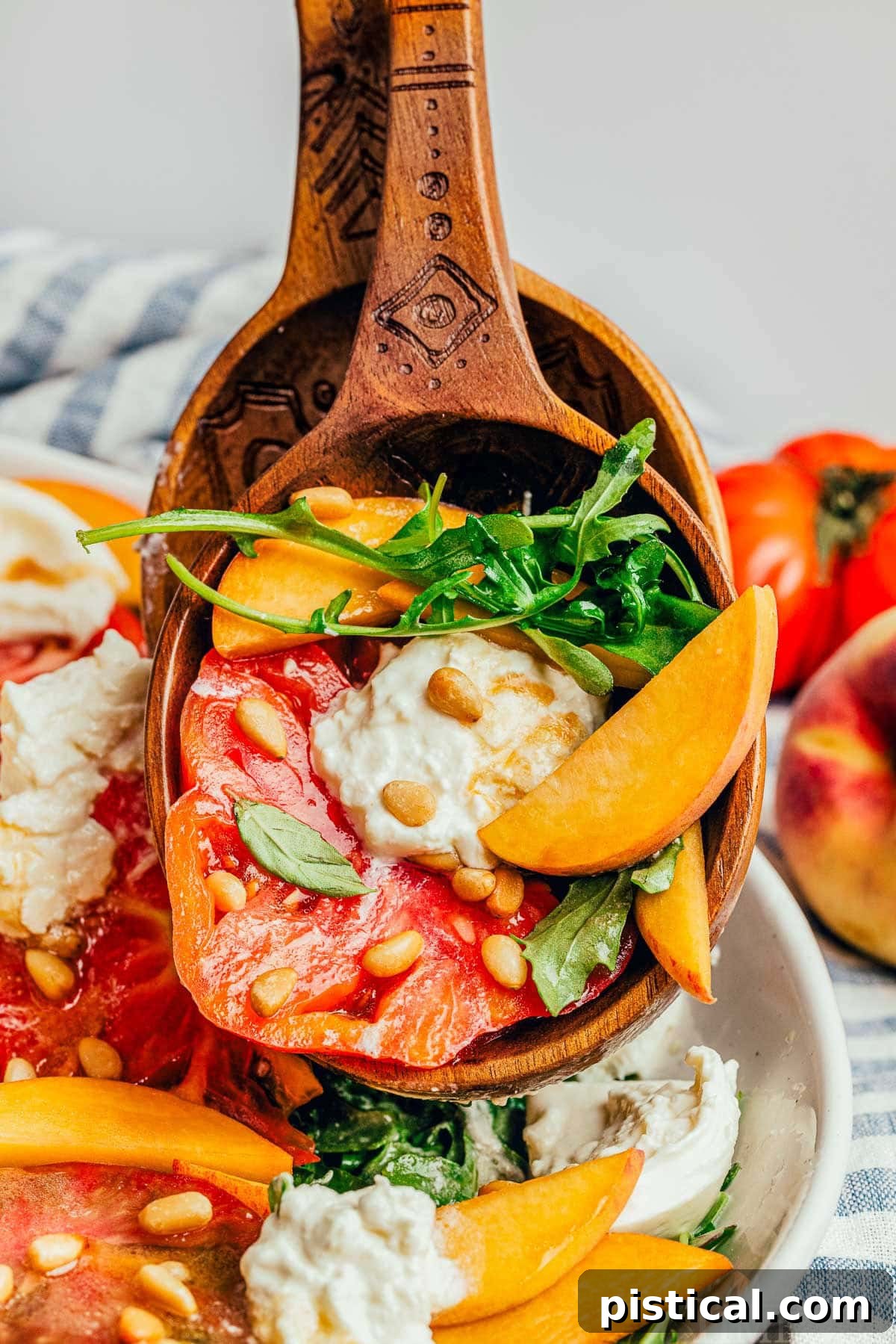 A pair of salad tongs lifting a generous scoop of Peach Burrata Salad, showcasing the vibrant combination of peaches, burrata, heirloom tomatoes, and arugula with toasted pine nuts.