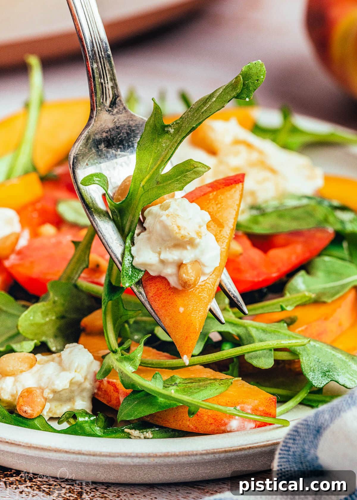 A fork holding a perfect bite of Peach Burrata Salad, showcasing a piece of peach, burrata, tomato, and arugula, poised above a full plate of the salad.