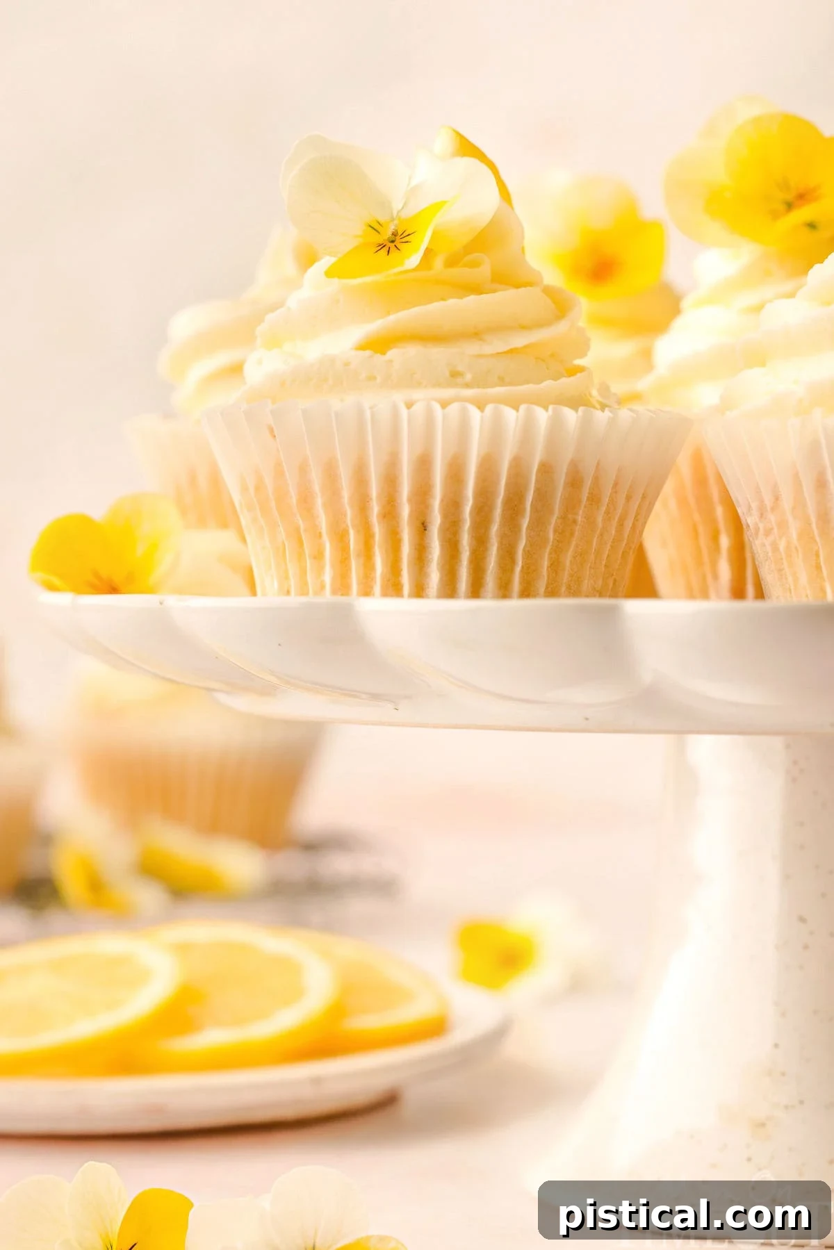 Zesty Lemon Delight Cupcakes 4 Lemon cupcakes on a scalloped edge white cake stand. Lemon slices and more cupcakes can be seen in the background.