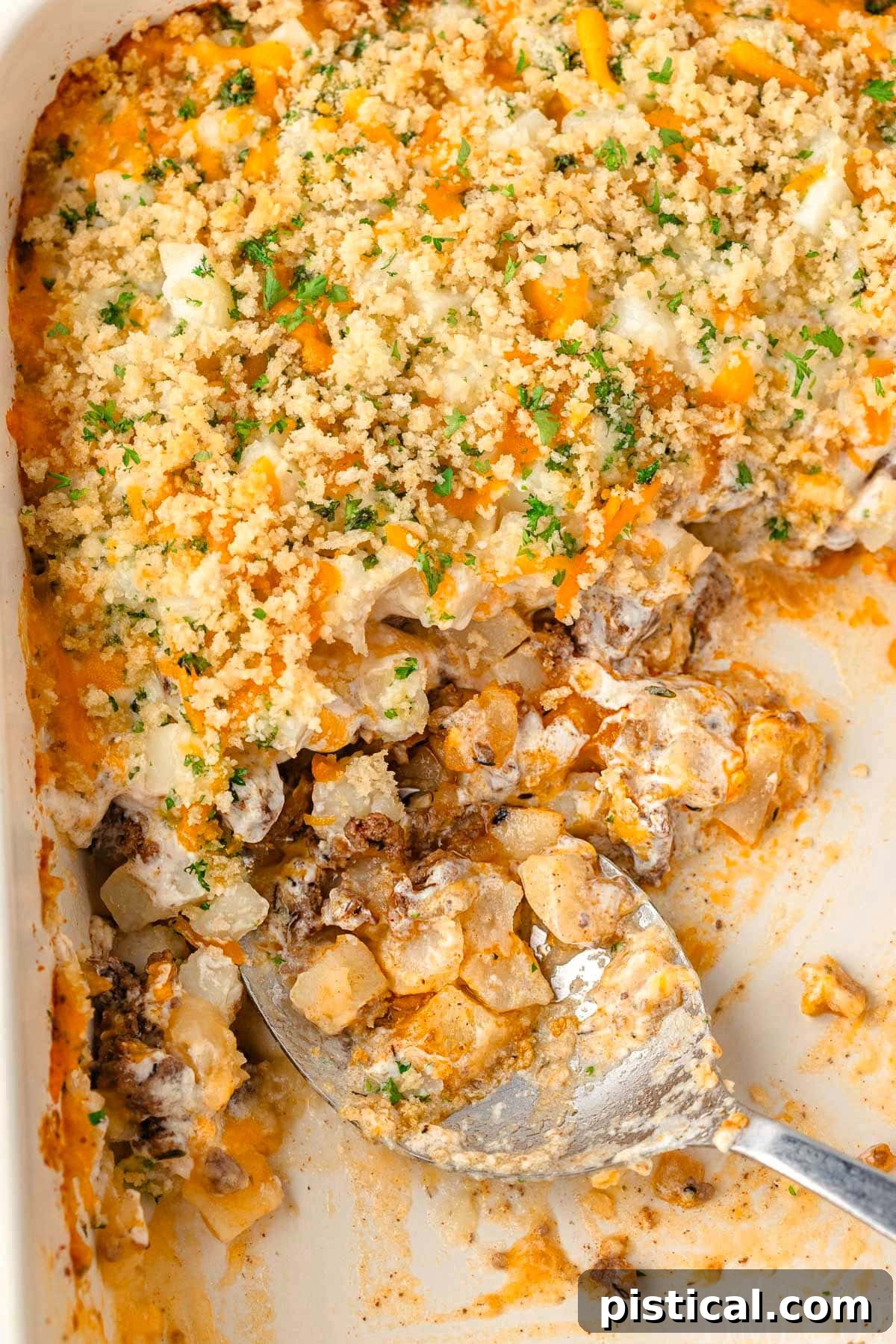 Ground beef casserole with a large serving spoon in a casserole dish, showing layers of beef, potatoes, and cheese.