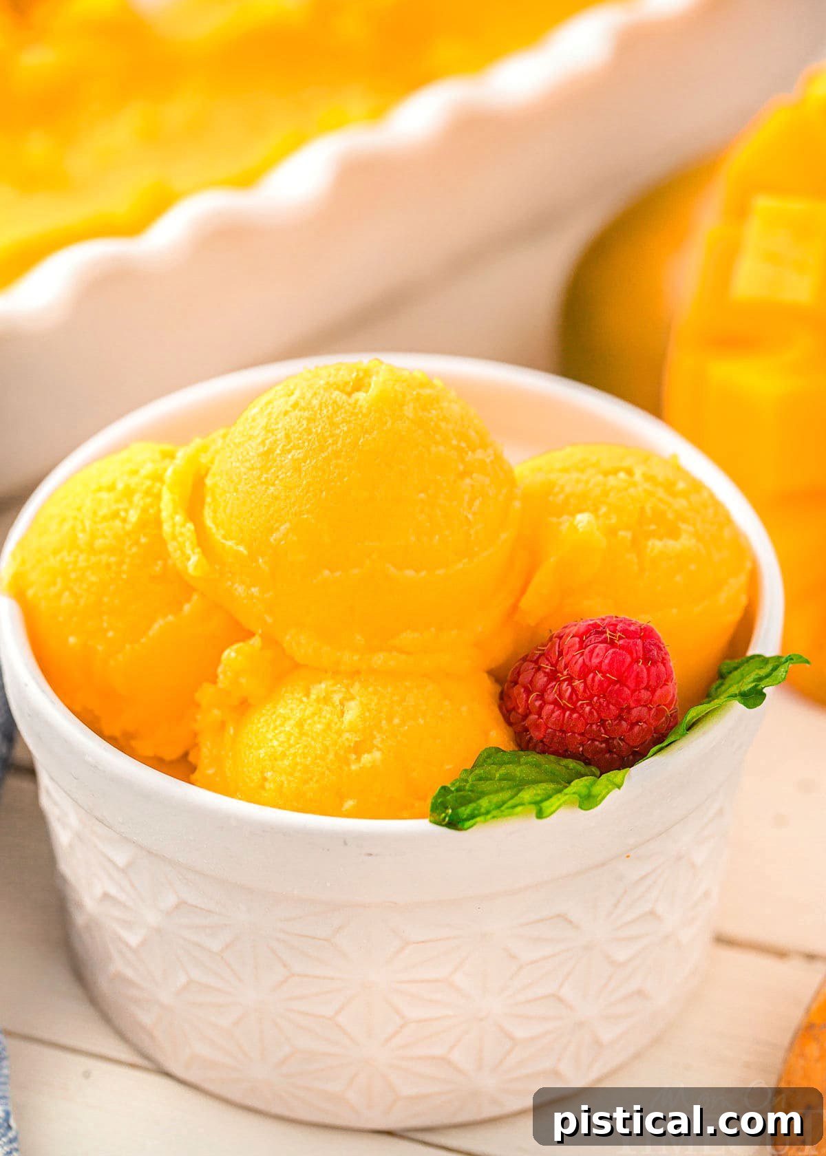Close-up of scoops of bright yellow mango sorbet in a white bowl, garnished with fresh mint.