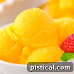 Scoops of mango sorbet in a white bowl topped with fresh mint and raspberries.