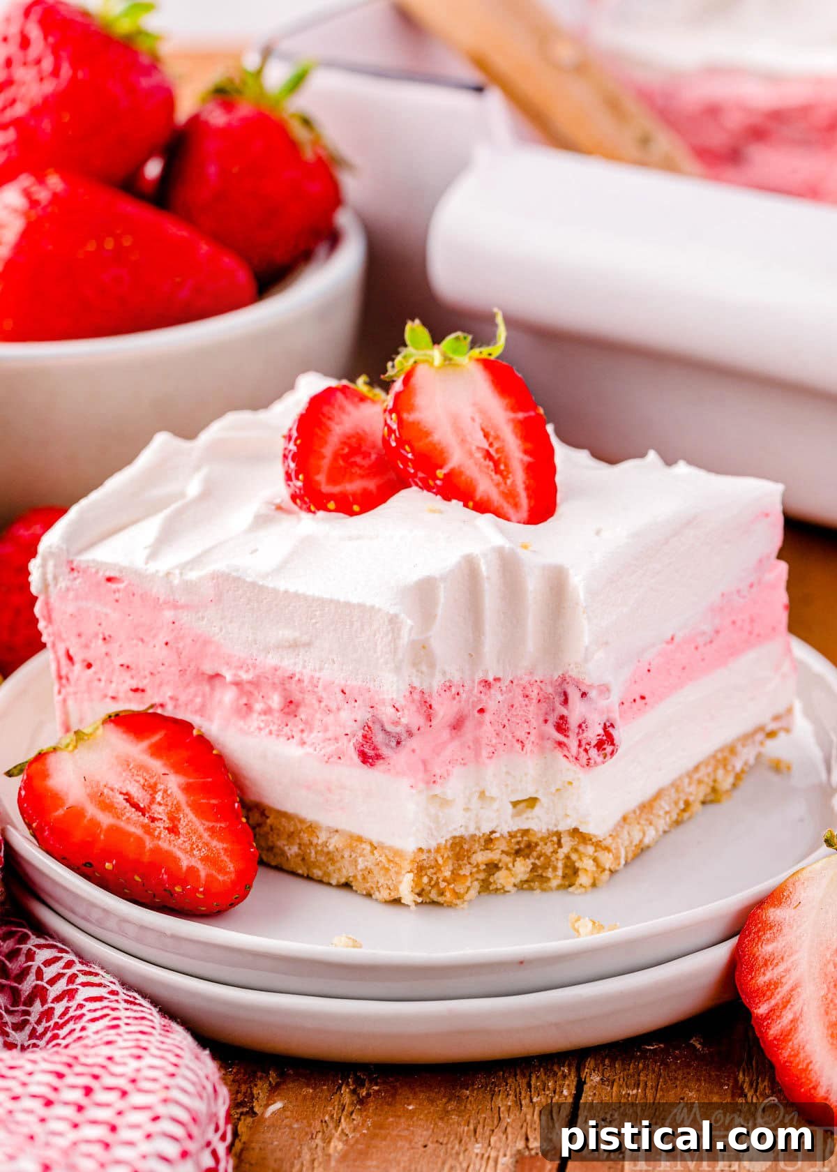 Layered Strawberry Dream 2 A square of strawberry dessert lasagna on a round white plate topped with whipped cream and fresh strawberries. A bowl of strawberries can be seen in the background.