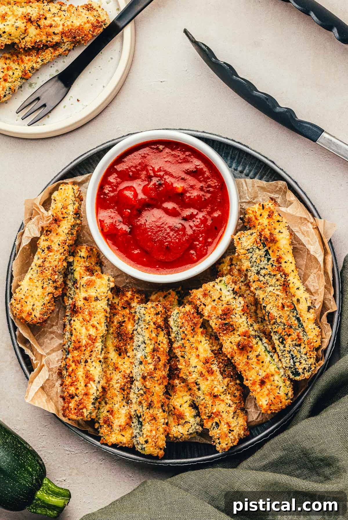 Top down view of air fryer zucchini on a metal plate with a small bowl of marinara to dip the fries in. Crispy golden zucchini fries are arranged around a vibrant red marinara sauce, showcasing their perfectly cooked texture and appetizing appearance.