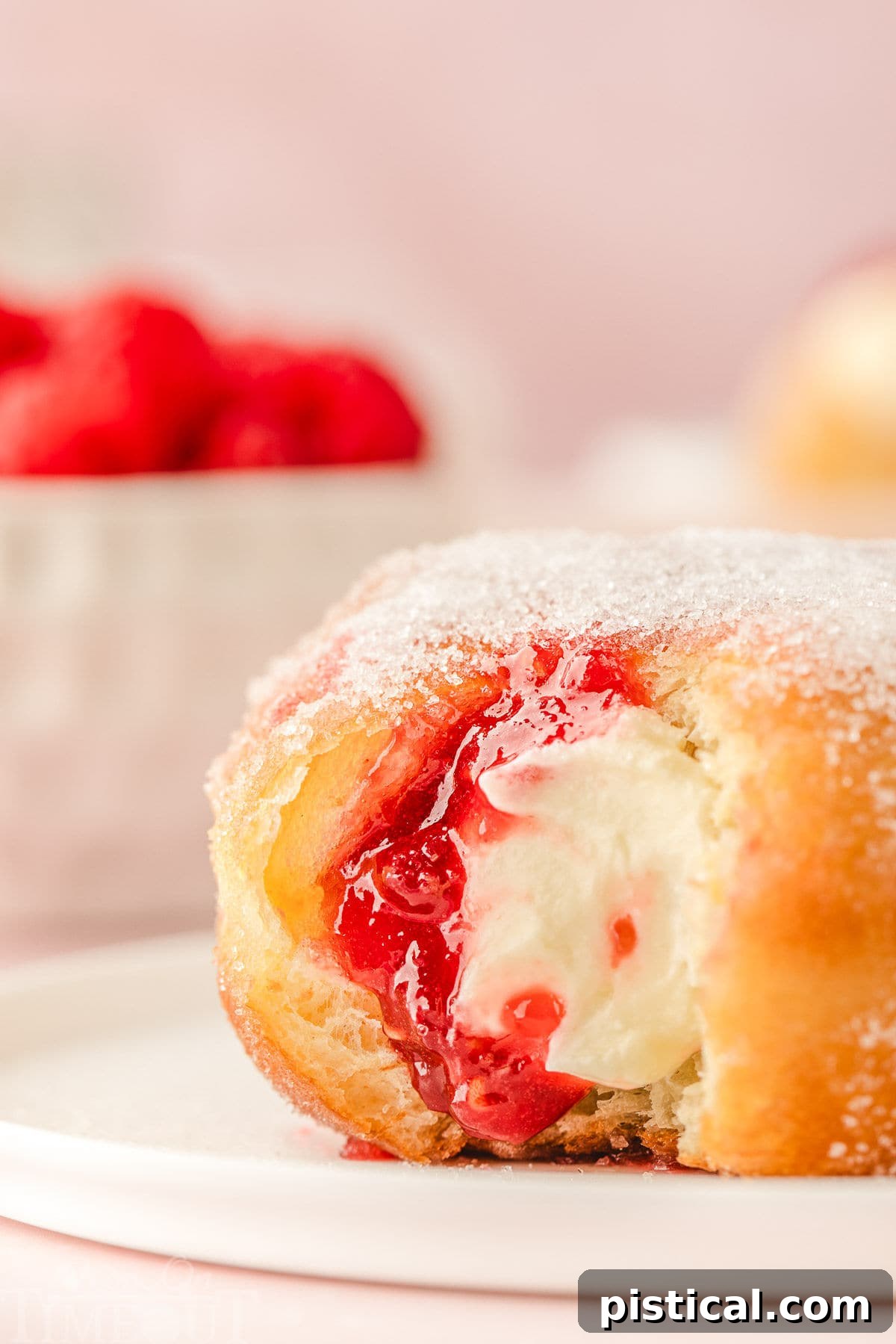 A single raspberry cheesecake stuffed donut on a small round white plate, showing a bite taken out of it to reveal the creamy filling.
