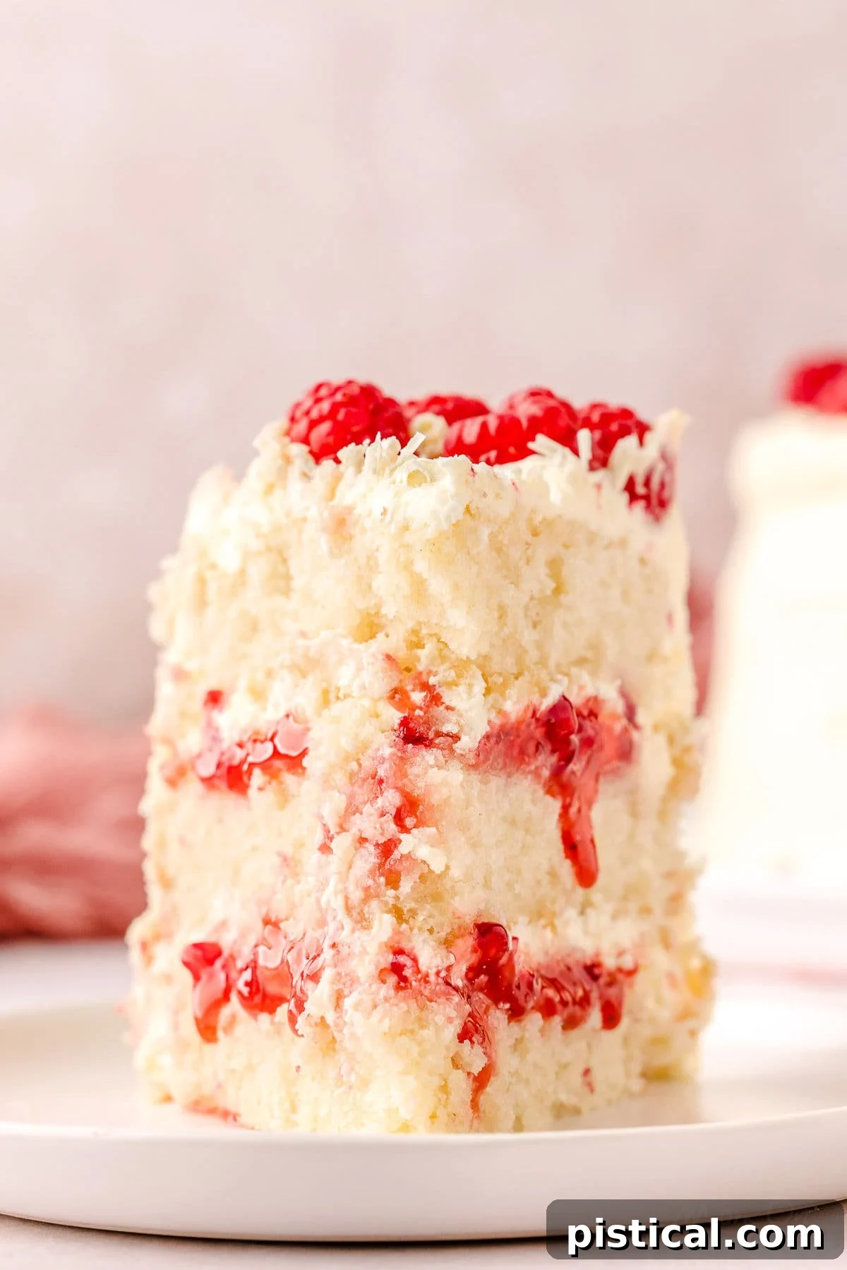 A perfectly cut slice of White Chocolate Raspberry Cake on a white round plate, adorned with fresh raspberries and delicate white chocolate curls, showcasing its beautiful layers.