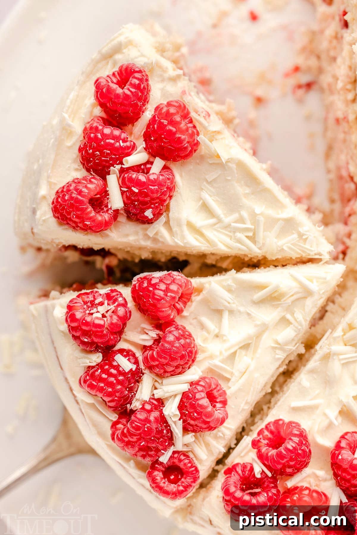 An overhead view of a White Chocolate Raspberry Cake, already cut into generous slices, revealing its stunning layers of cake, raspberry, and frosting. The cake is beautifully garnished with fresh red raspberries and white chocolate shavings.