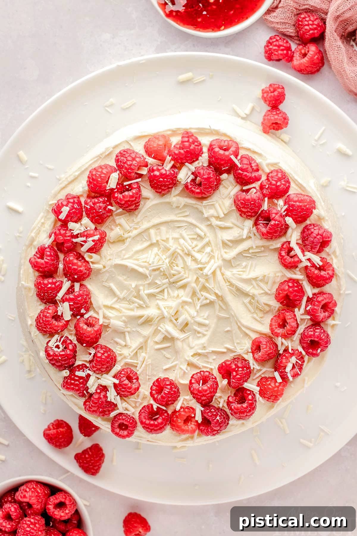 An inviting top-down view of a whole White Chocolate Raspberry Cake, elegantly presented on a large white cake plate, ready to be sliced and served.