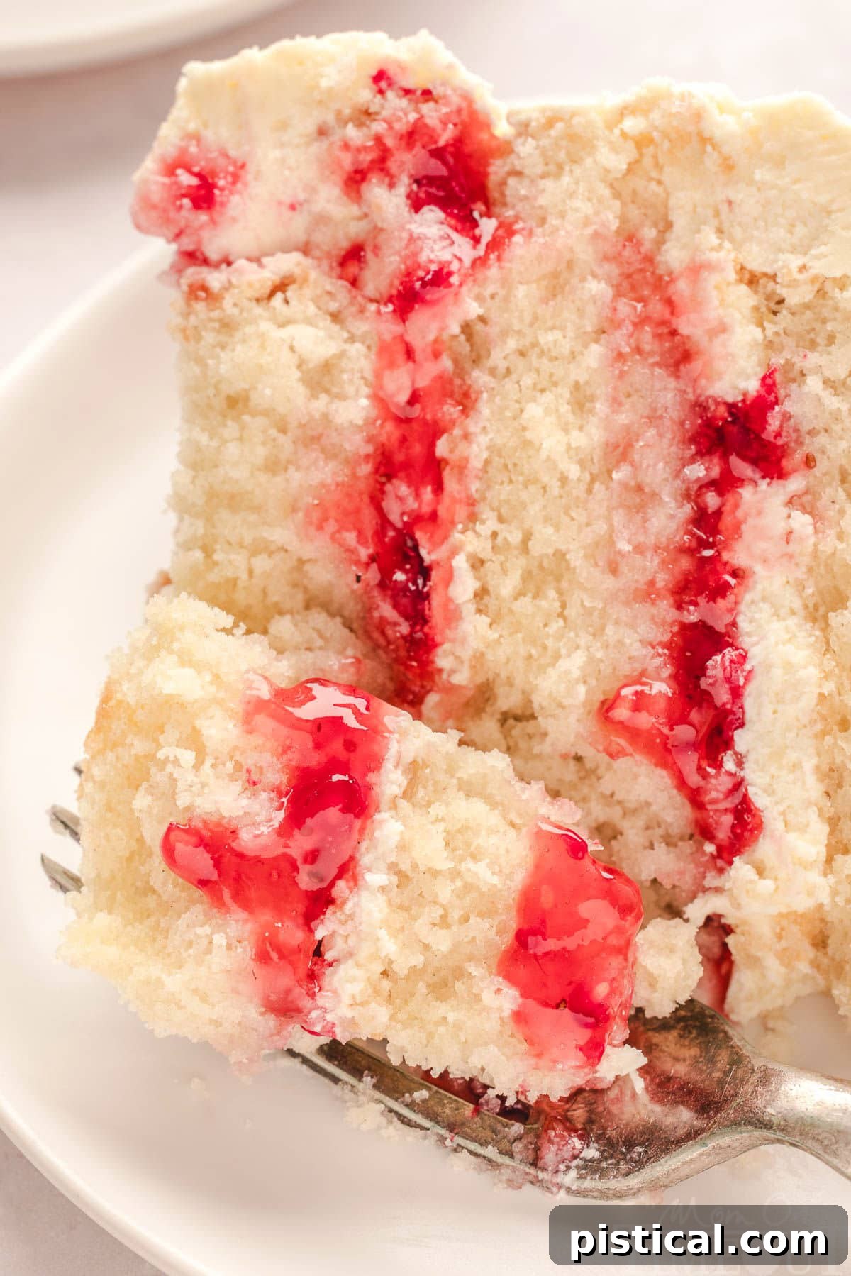 A close-up of a slice of Raspberry White Chocolate Cake on a white round plate, with a fork poised to take a bite, highlighting its tempting layers and perfect texture.