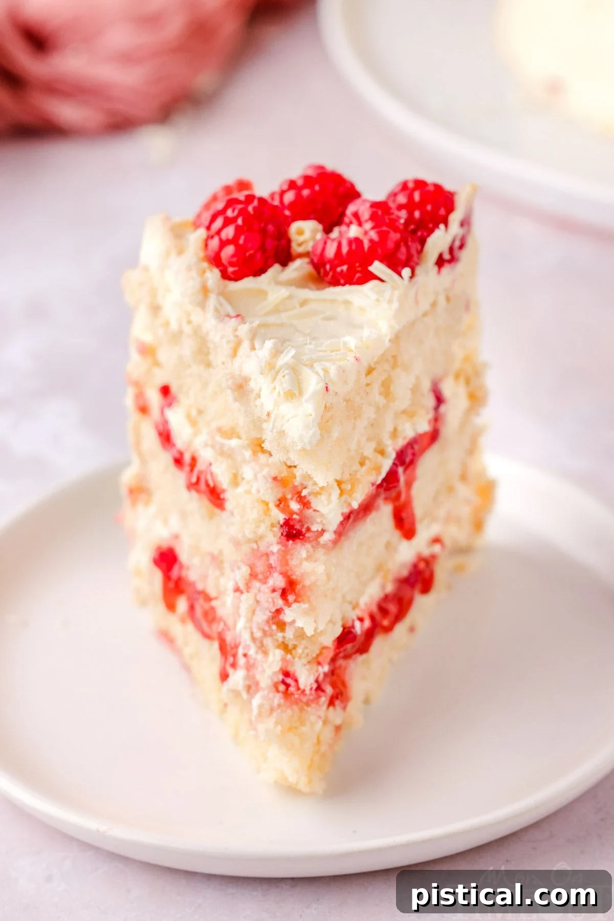 A beautiful slice of White Chocolate Raspberry Cake on a white round plate, expertly topped with fresh raspberries and decorative white chocolate curls, ready to be enjoyed.