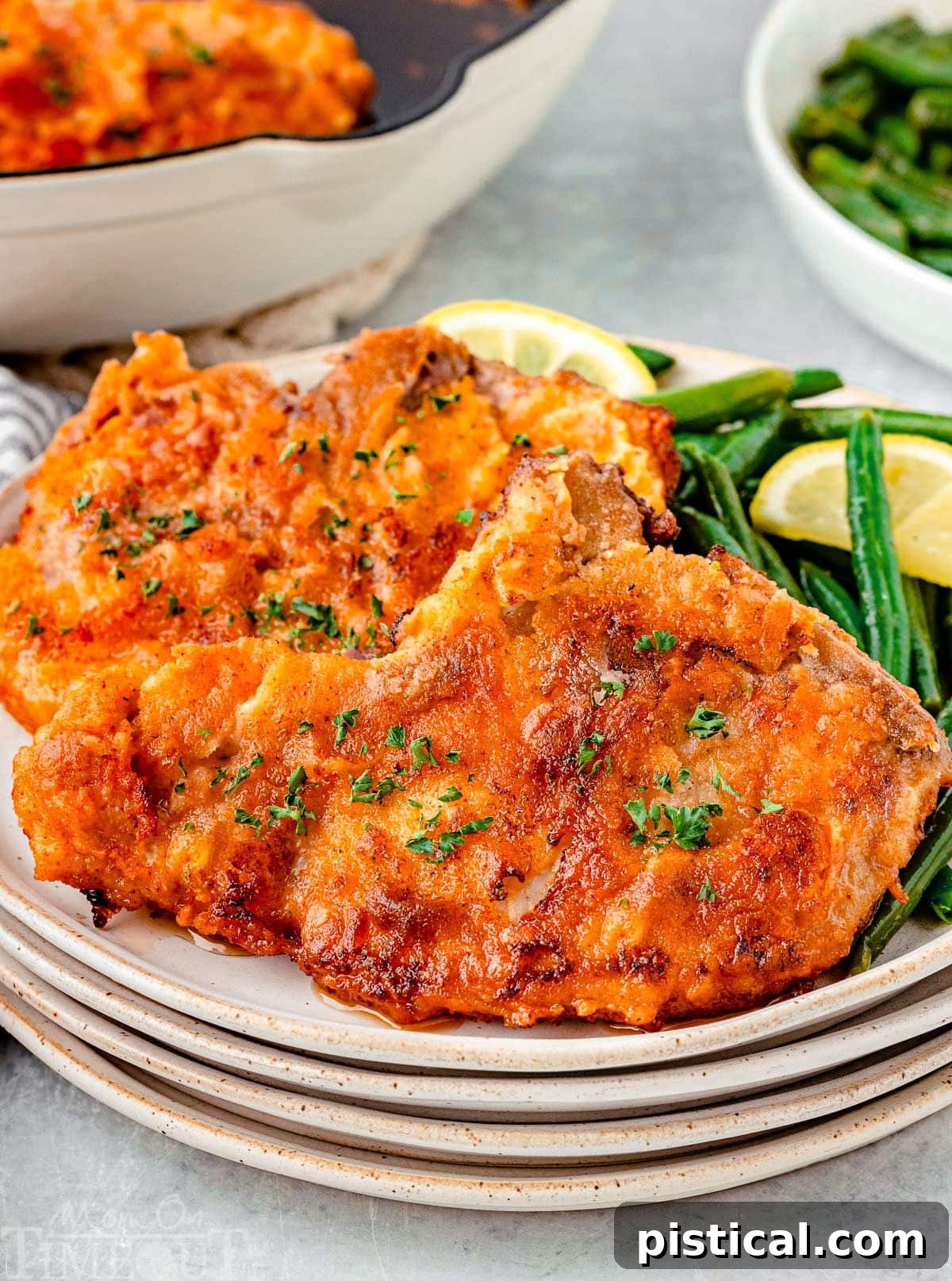 Two golden-brown fried pork chops resting on a stack of small, rustic plates, accompanied by vibrant green beans and a bright lemon wedge, ready to be enjoyed as a hearty meal. The crispy exterior of the pork chops is visibly appealing.