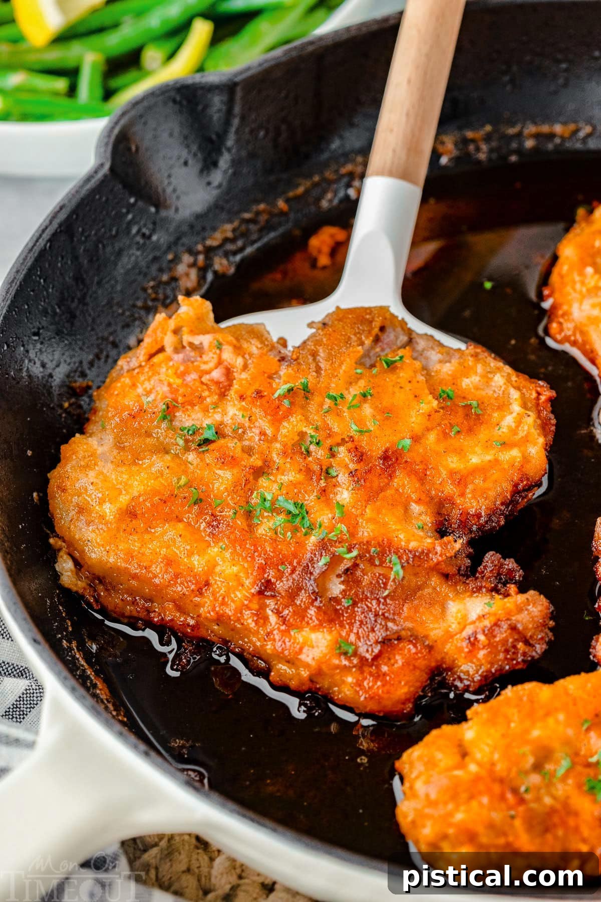 A kitchen spatula carefully lifting a perfectly fried pork chop from a sizzling cast iron skillet, showcasing its golden-brown, crispy crust. The hot oil glistens around the remaining chops.