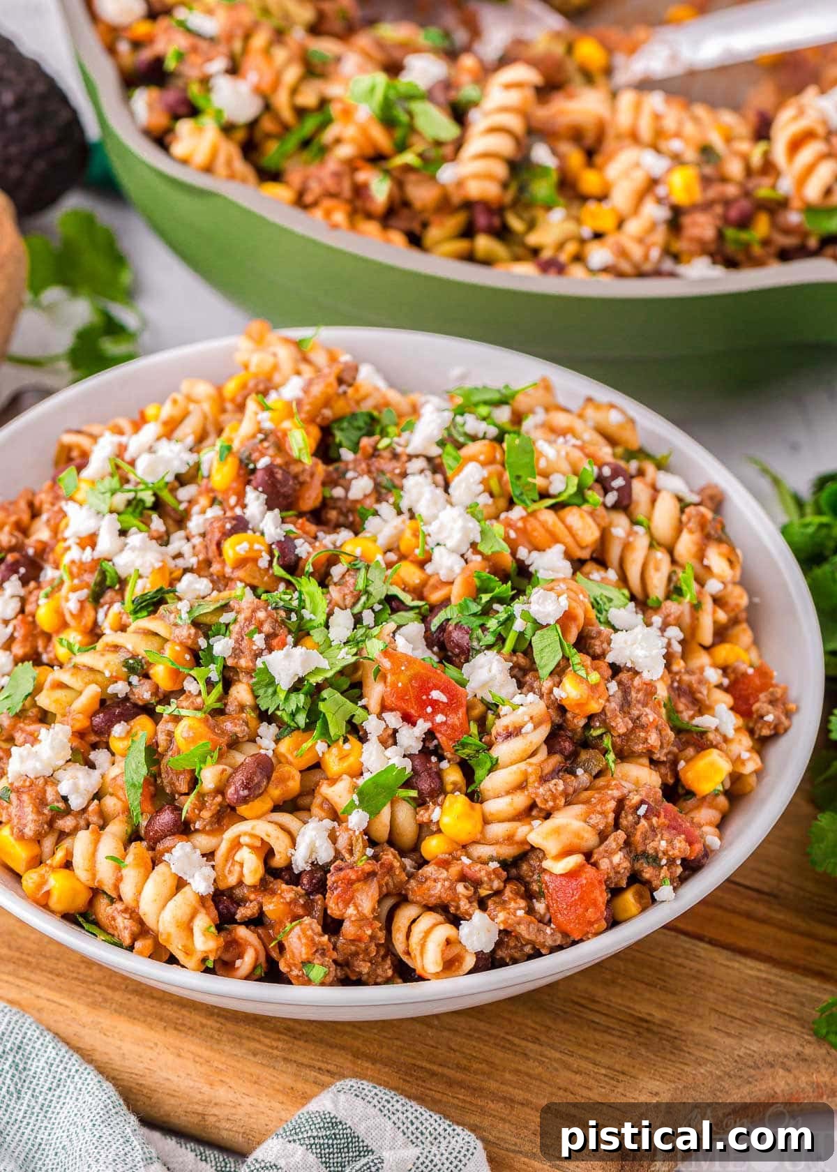Tex-Mex Pasta Skillet 2 Angled view of a bowl filled with vibrant taco pasta, with a green skillet containing more pasta in the soft background. The dish is garnished with fresh cilantro and a sprinkle of cheese, highlighting its delicious appeal.