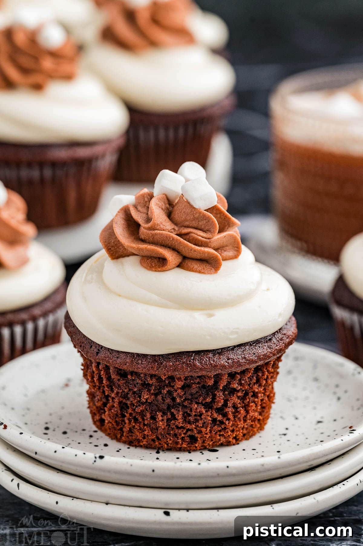 Velvet Hot Chocolate Cupcakes 2 A hot chocolate cupcake on a stack of white plates with more cupcakes in the background. The cupcake is generously frosted with marshmallow and topped with whipped cream and mini marshmallows.