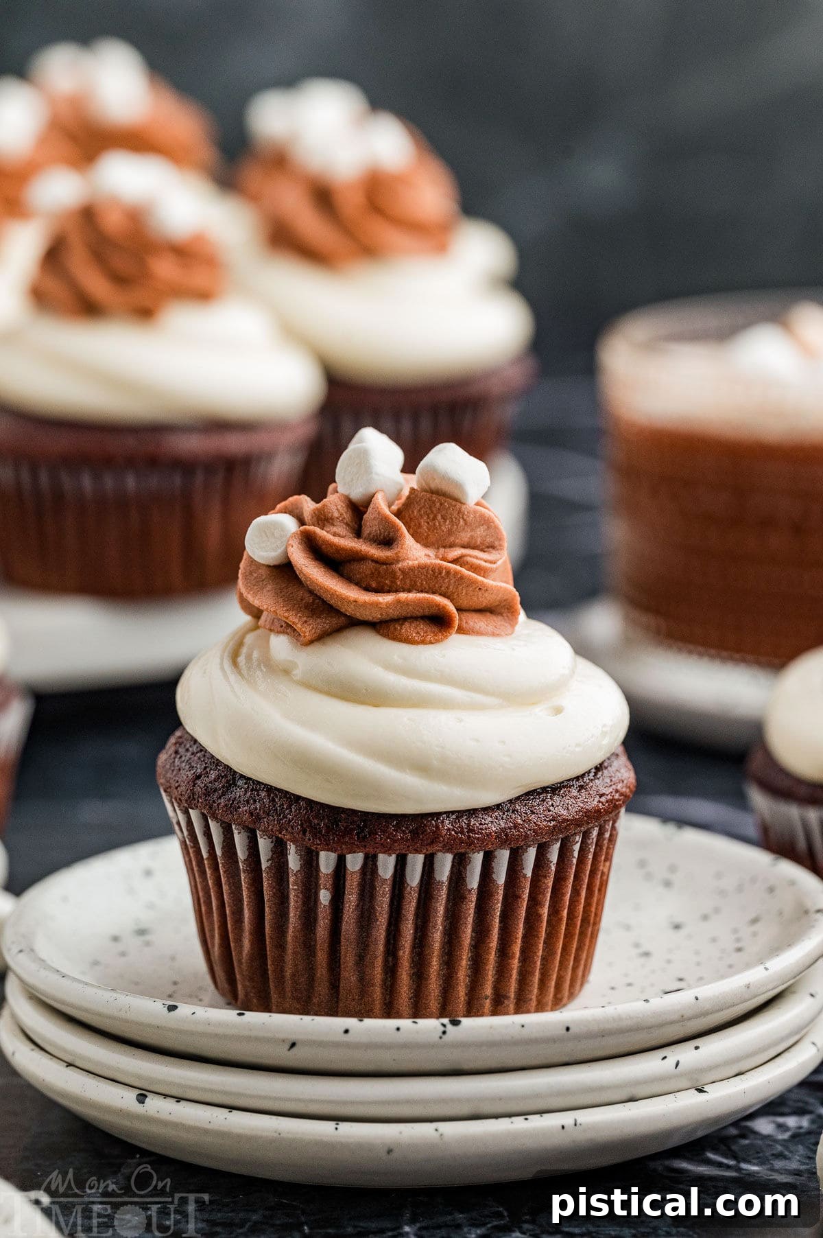 Velvet Hot Chocolate Cupcakes 10 A hot chocolate cupcake on a stack of white plates with more cupcakes in the background, showcasing its rich frosting and toppings.