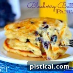 A stack of Blueberry Buttermilk Pancakes on a white plate with syrup on a blue placemat