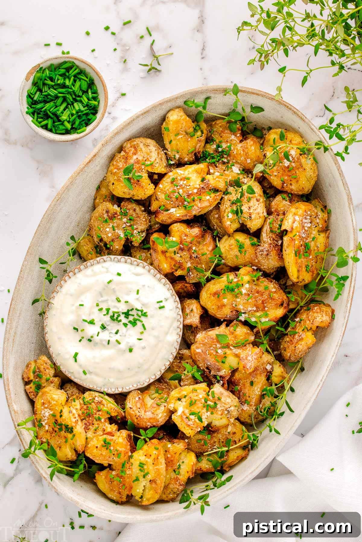Top down view of an oval platter of smashed potatoes garnished with thyme and chives. A small bowl of dip is nestled amongst the smashed potatoes.