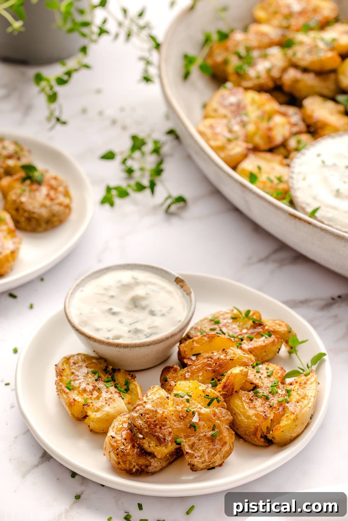 Smashed potatoes plated on a small round white plate with sour cream dip. Platter with more smashed potatoes in the background.