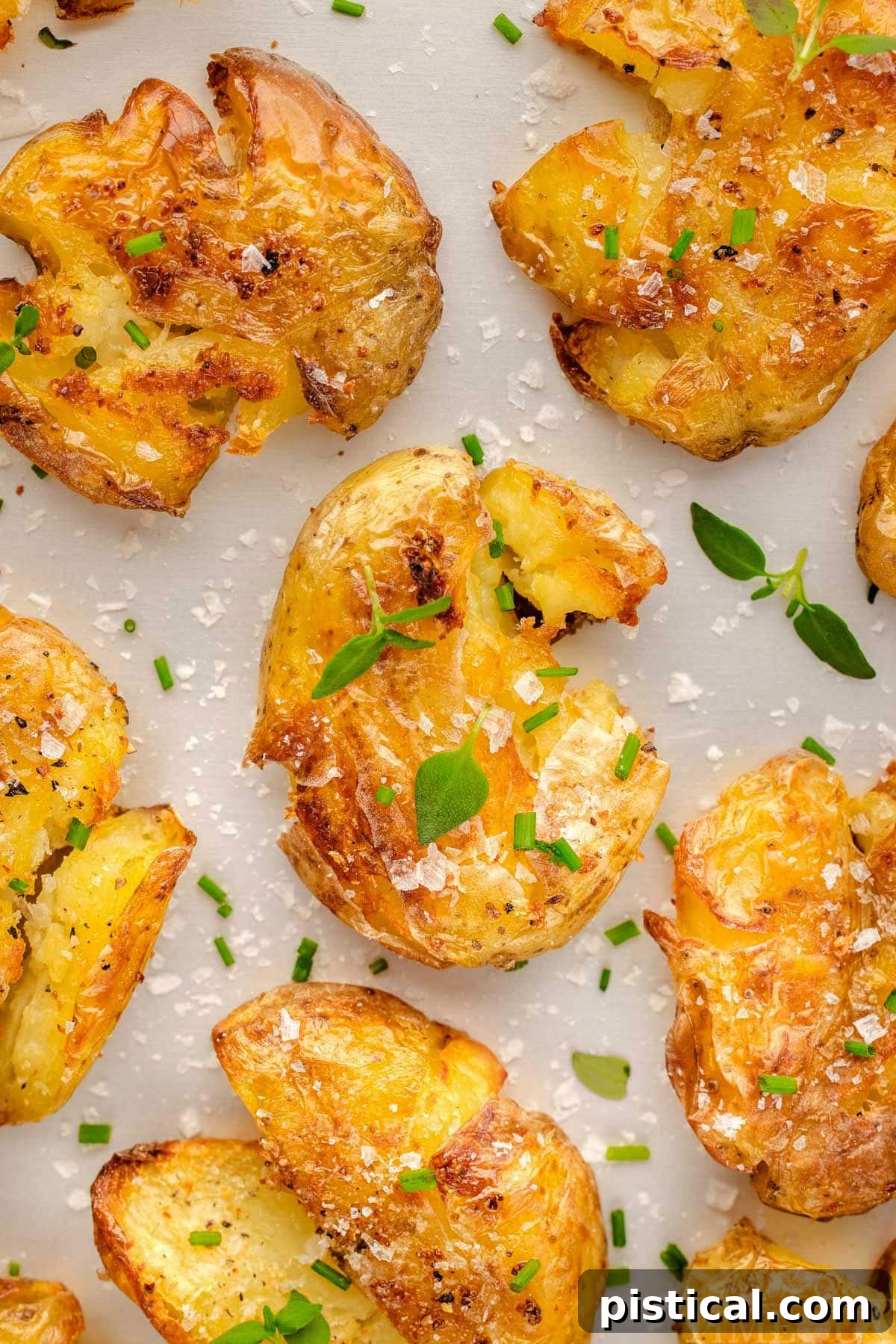Top down view of smashed potatoes recipe on a white surface. The potatoes have been seasoned with salt, pepper and are topped with chives and thyme.