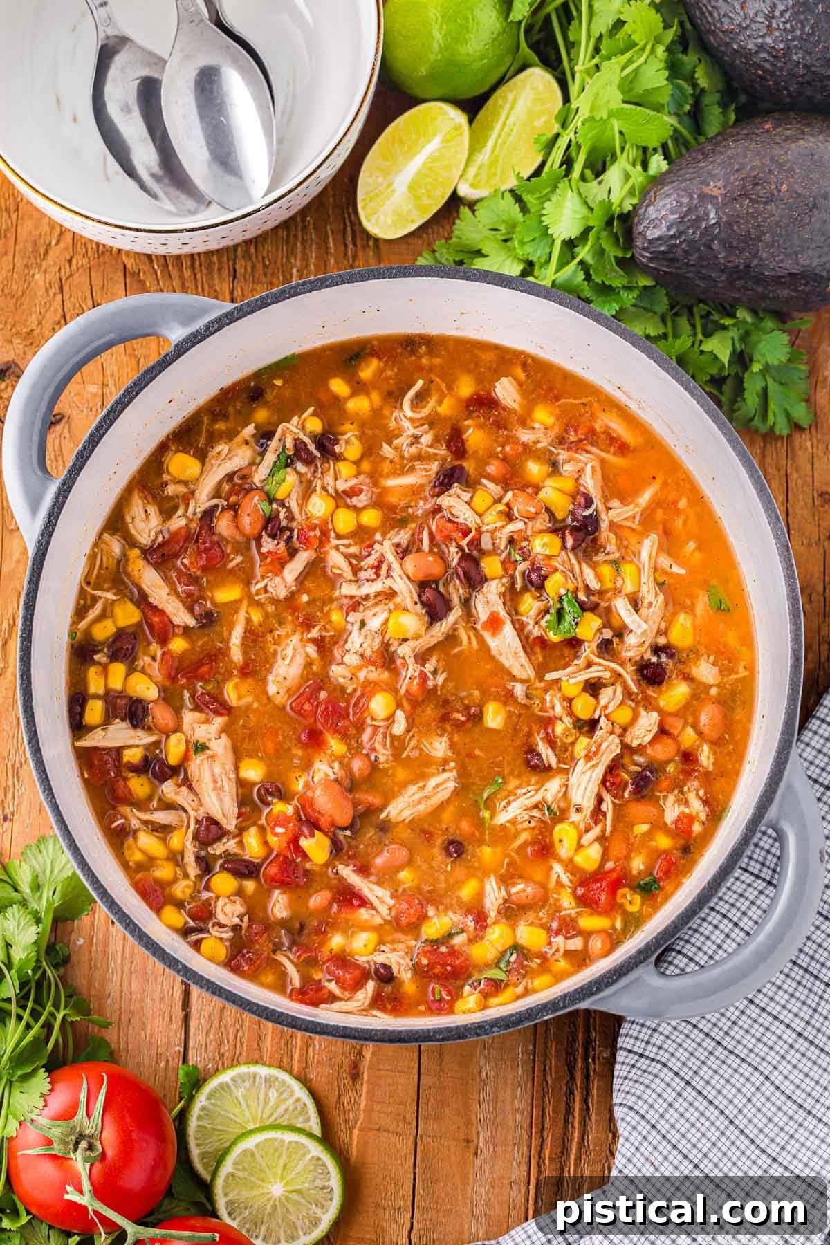 A top-down view of chicken taco soup simmering in a Dutch oven, surrounded by fresh garnishes and empty bowls on a rustic table.