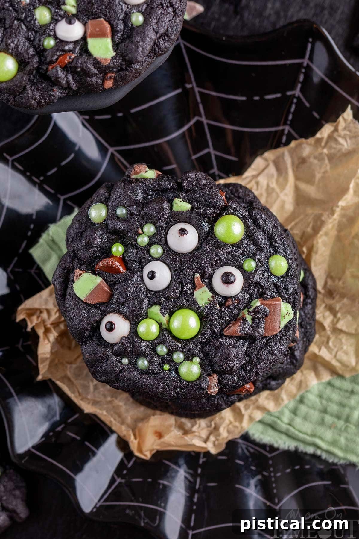 Mint Chocolate Chip Monster Halloween Cookies stacked on brown parchment paper and a black web design plate. Cookies feature playful candy eyes and luminous green sugar pearls for decoration.