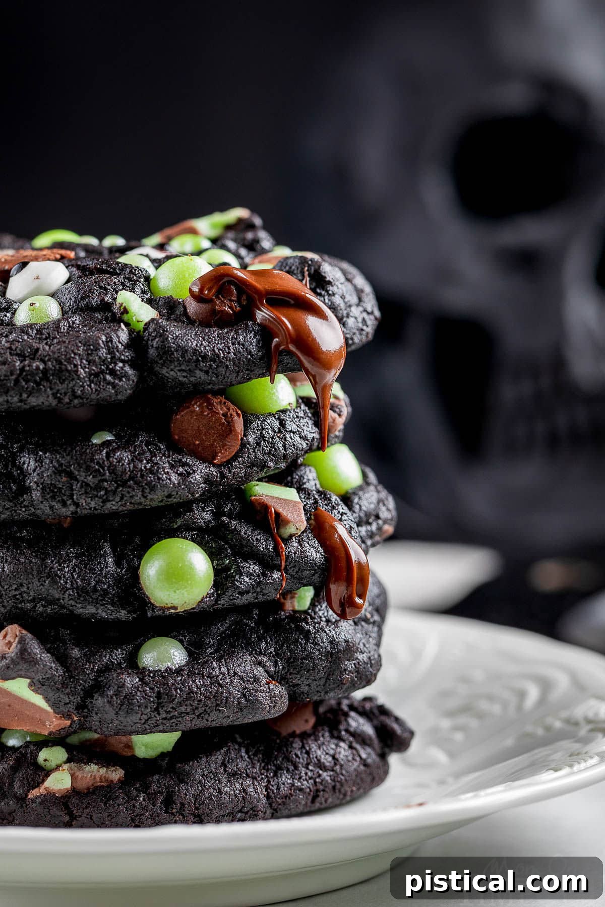Close-up side view of a stack of Halloween monster cookies. These dark black cookies, made with black cocoa powder, are adorned with melting dark chocolate chips and festive green pearl candies, creating a delightful texture and appearance.