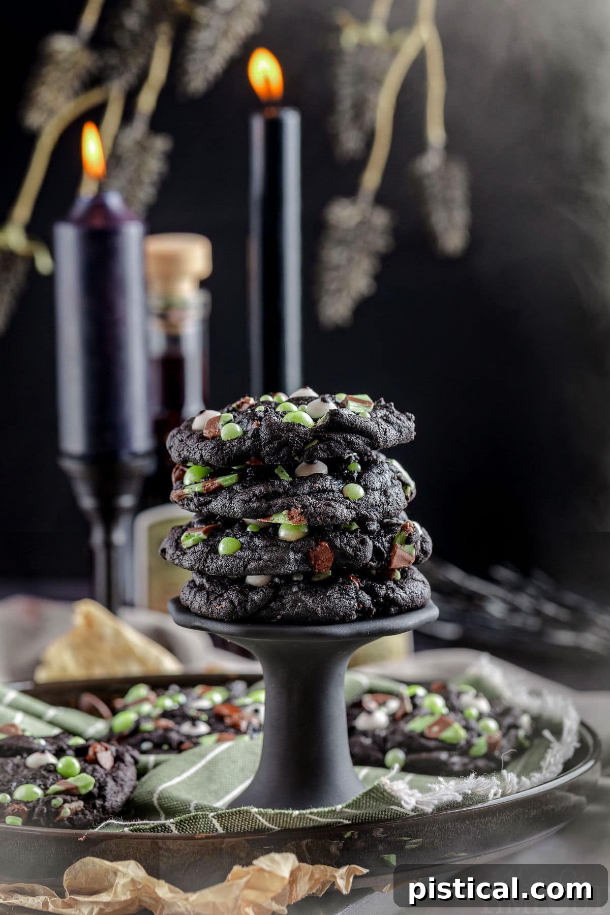 A towering stack of Mint Chocolate Chip Monster Halloween Cookies, elegantly presented on a black stand. The background features atmospheric black candles and shadowy branches, setting a spooky and festive scene.