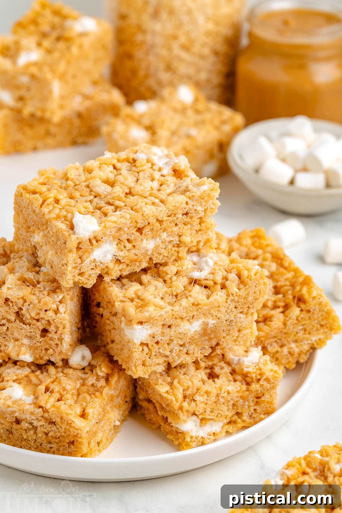 Peanut butter rice krispie treats stacked on a white round plate with more treats in the background, showcasing their gooey texture.