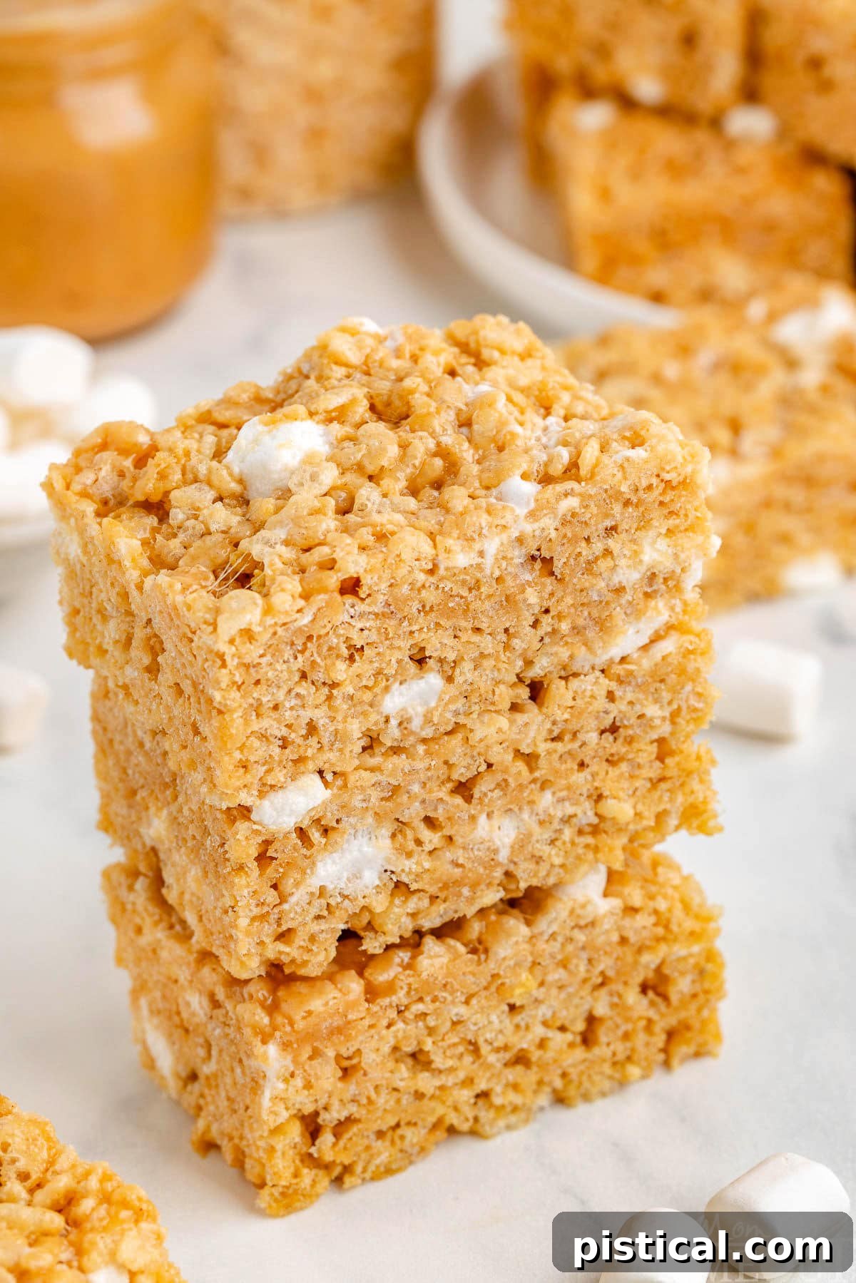 Peanut butter rice krispie treats stacked three high on white surface with marshmallows scattered about, highlighting their appealing appearance.