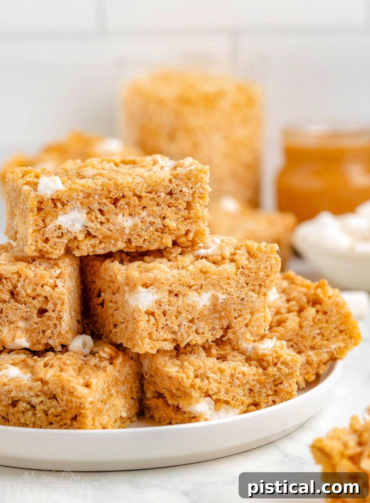 Peanut butter rice krispie treats stacked on a white round plate with more treats in the background, inviting a taste.