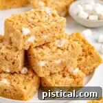 Peanut butter rice krispie treats stacked on a white round plate with more treats in the background.