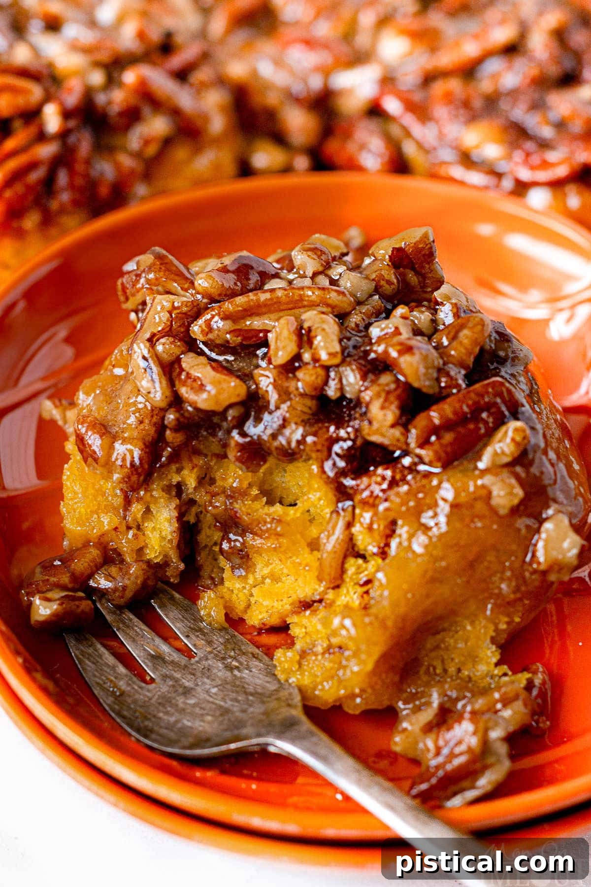A single, perfectly baked pumpkin sticky bun rests on a vibrant orange plate, a fork nearby, with one tempting bite already taken.