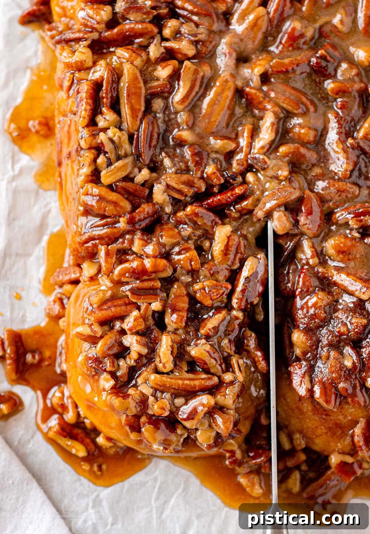 An overhead view captures a single pumpkin sticky bun being carefully sliced away from the cluster of remaining buns on a parchment-lined cutting board, highlighting its individual perfection.
