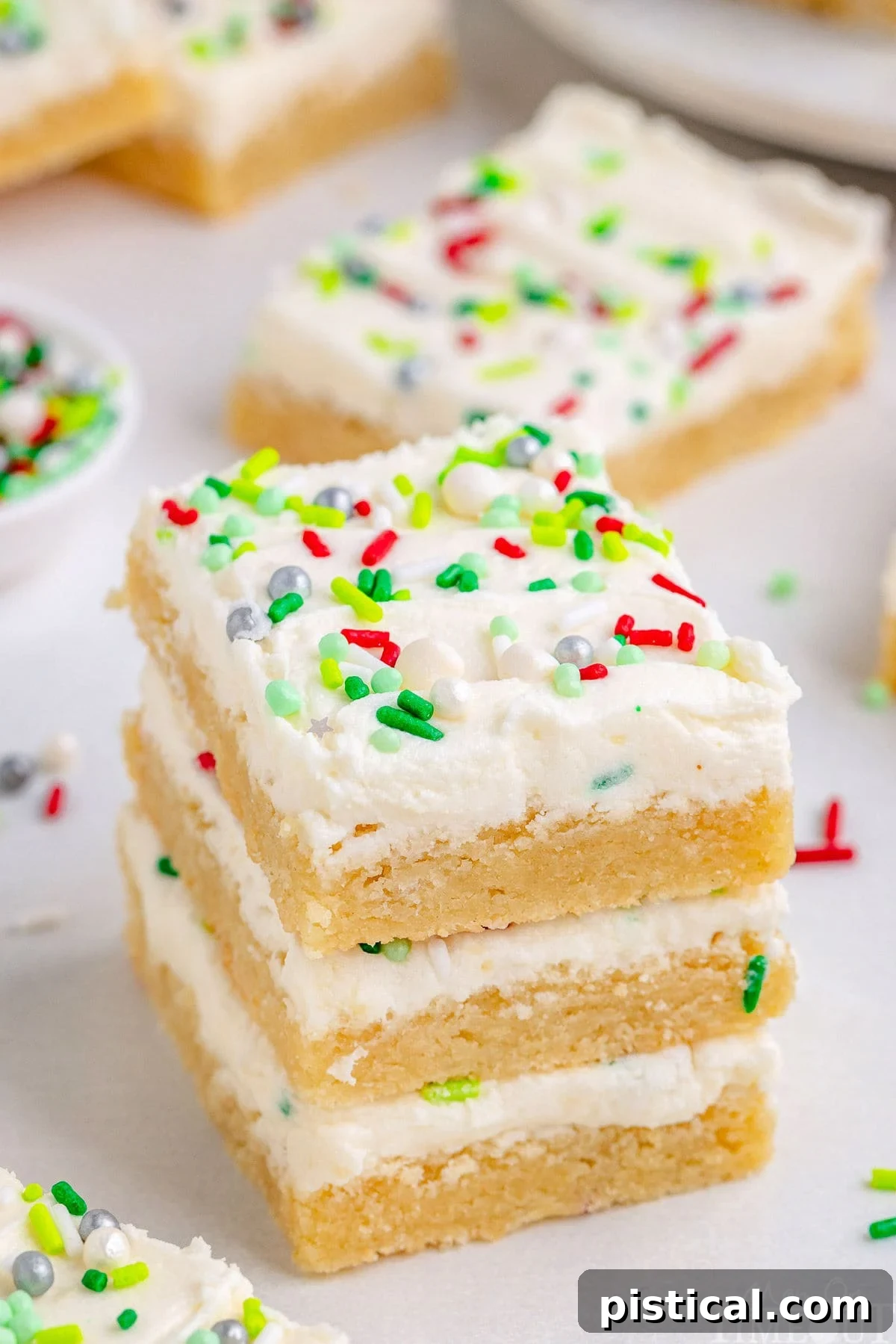 A stack of three soft, frosted Christmas Sugar Cookie Bars, with more beautifully decorated bars visible in the background, showing off their festive appeal.