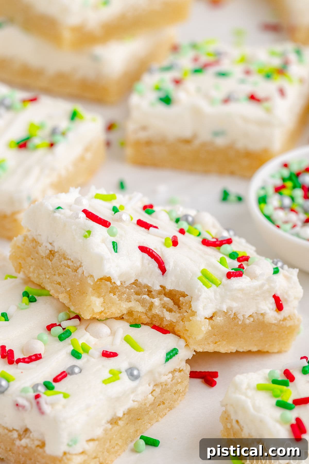 A close-up shot of a single Christmas Sugar Cookie Bar with a bite taken out, resting on another bar, showcasing its tender texture and creamy frosting. More bars are blurred in the background.