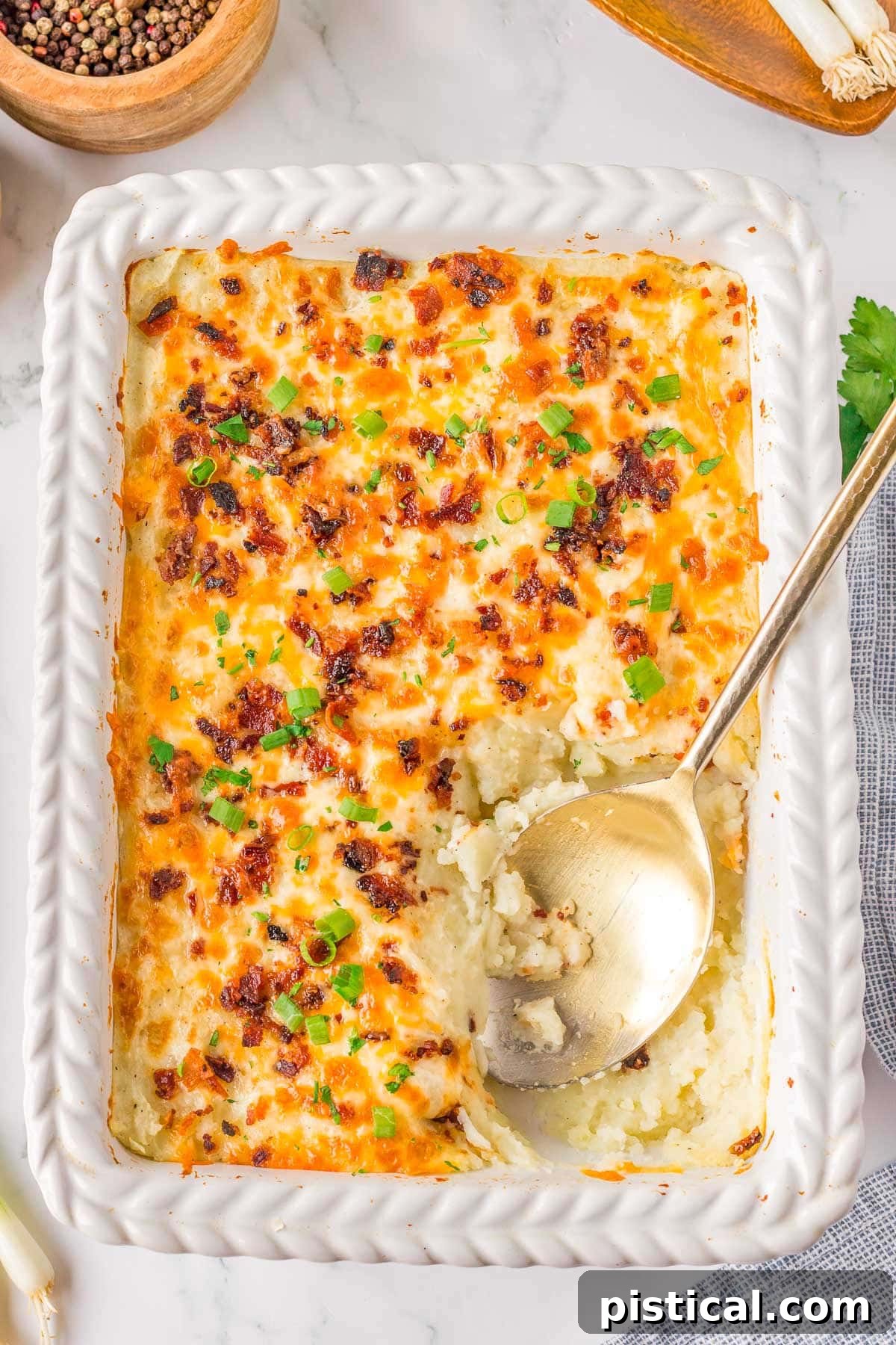 Velvety Potato Bake 3 Top down look at loaded mashed potato casserole in a white baking dish with a gold serving spoon resting in the dish where a serving has been removed.