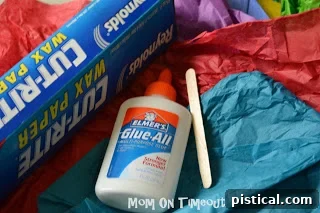 A child's hands carefully placing tissue paper pieces onto a glue-covered wax paper.