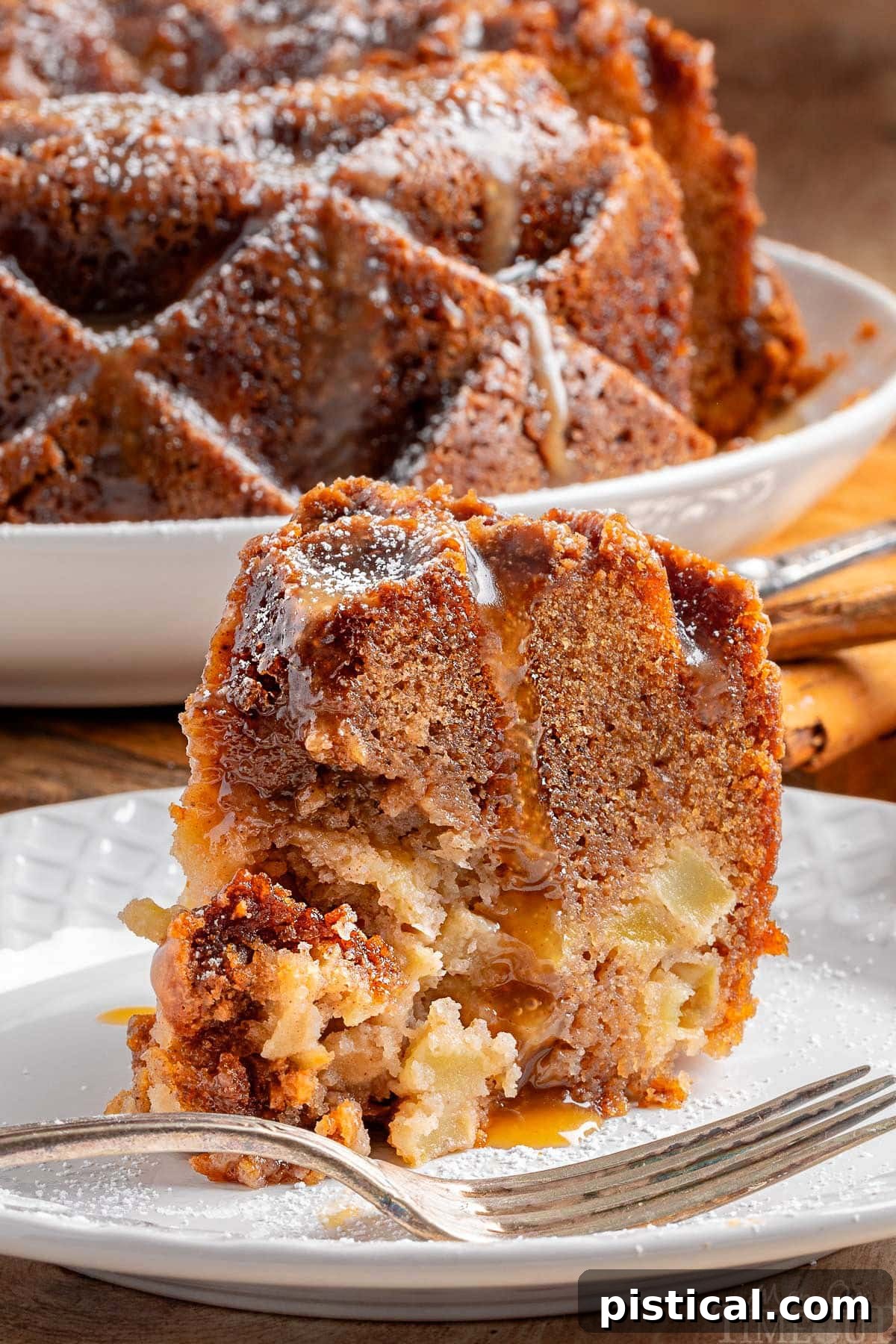A slice of Caramel Apple Bundt Cake with bourbon caramel glaze, sitting on a white plate, with the rest of the cake in the background.